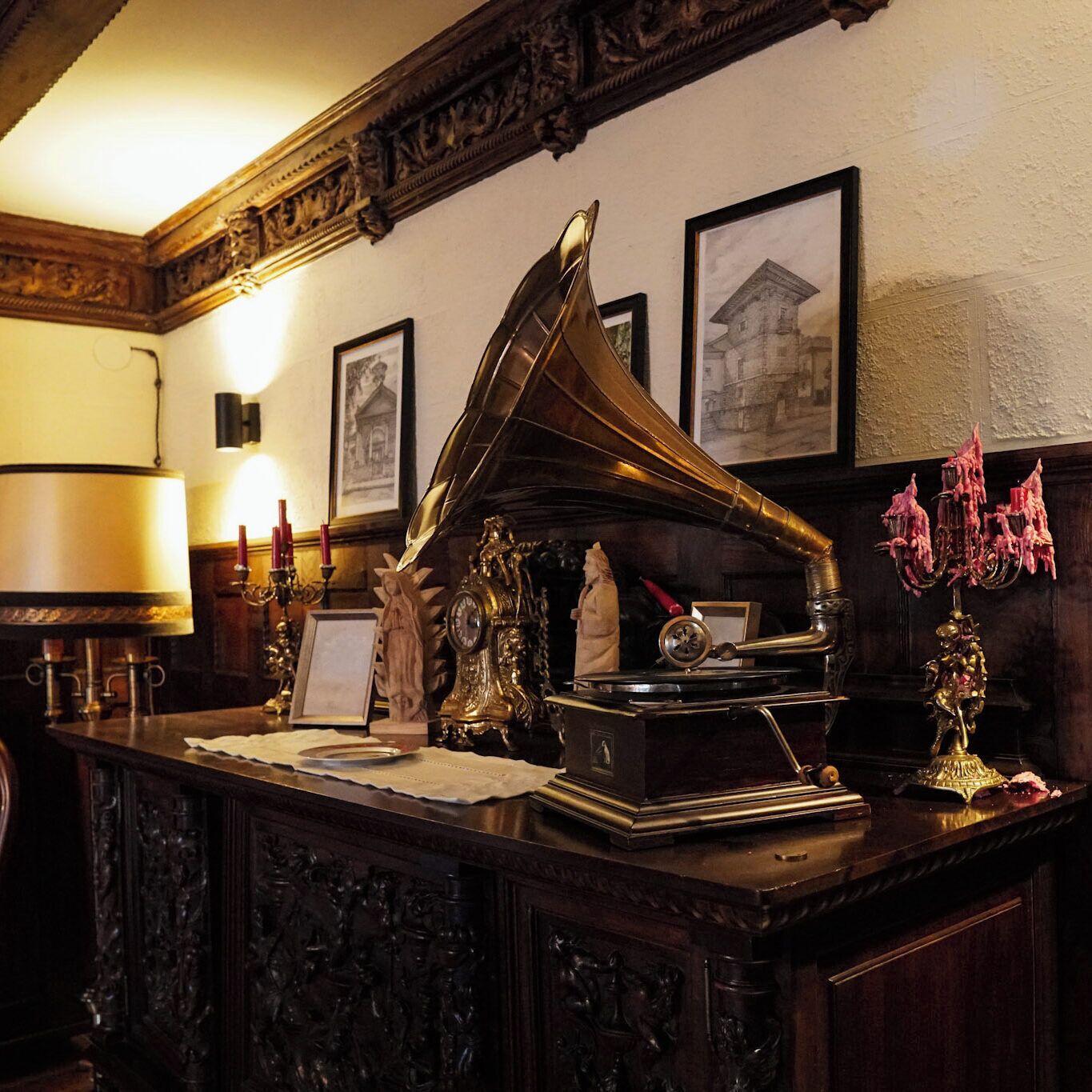 Un gramófono antiguo sobre un escritorio de madera con adornos labrados, un cuadro enmarcado en la pared y un candelabro con velas rosas.
