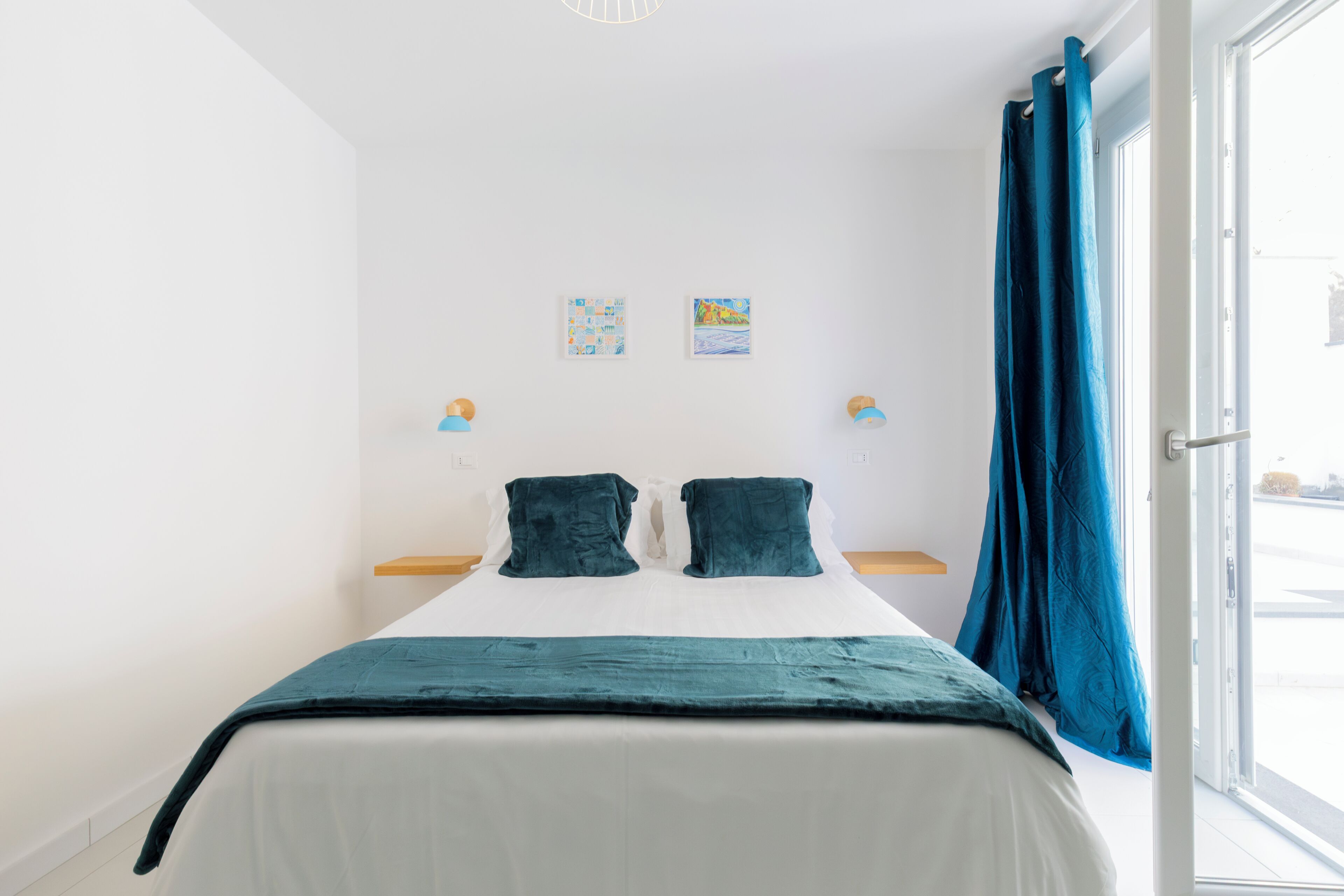 Una camera da letto con un letto, due cuscini, una coperta blu, una finestra con tende e due luci a parete.