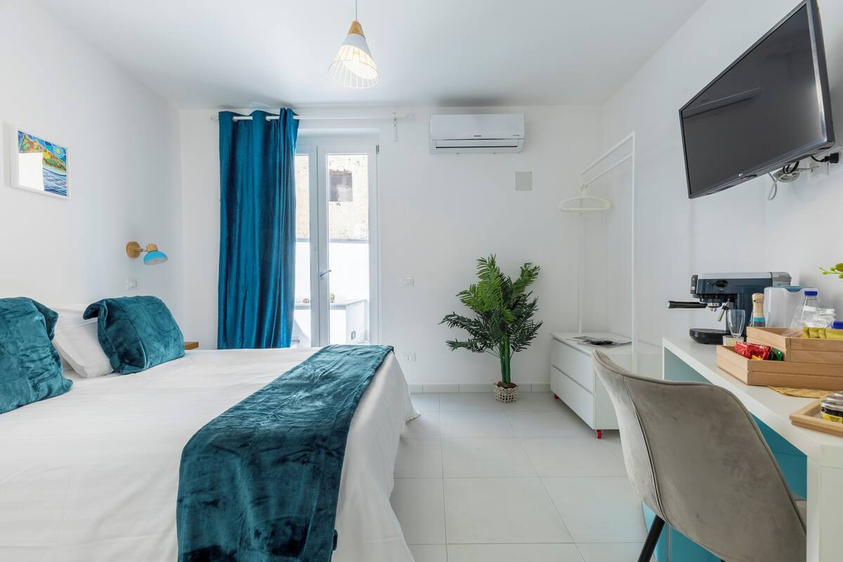 A modern hotel room with a bed, desk, chair, TV, air conditioning unit, and a potted plant.