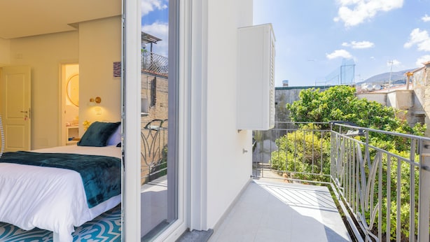 A hotel room with a bed, a small table with a lamp, and a balcony with a metal railing and greenery. - Via Roma Suites (Ischia)