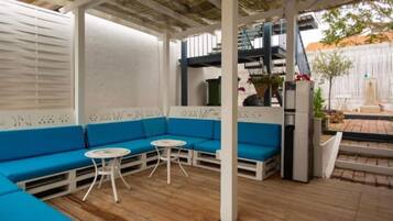 Un espace extérieur aménagé avec des meubles de jardin, comprenant des canapés bleus et des tables blanches, un sol en bois et une clôture blanche en arrière-plan.