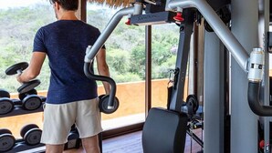 Un homme fait de l’exercice avec des haltères et une machine de musculation dans une salle de sport dotée d’un toit de chaume et de grandes fenêtres.