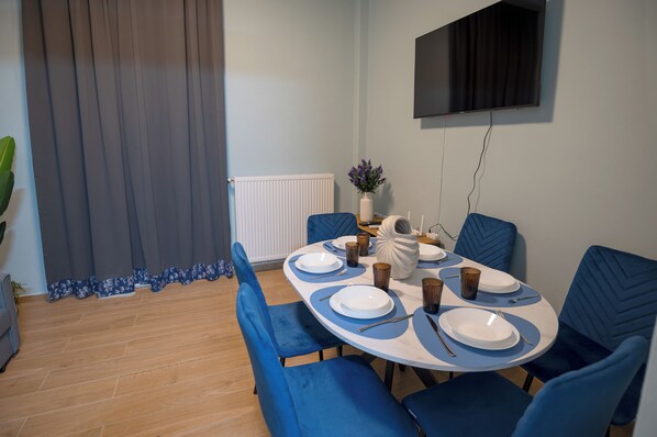 Un comedor con una mesa redonda dispuesta para cuatro personas, sillas tapizadas en azul, un jarrón blanco con flores moradas y un televisor de pantalla plana montado en la pared.