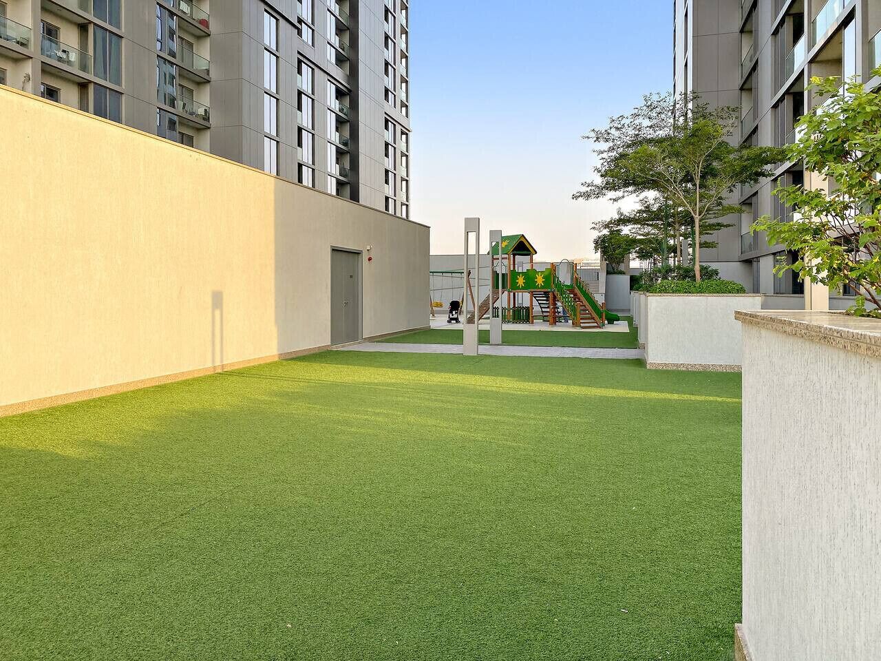 An outdoor area with artificial grass, a children's play structure, and a building with balconies.