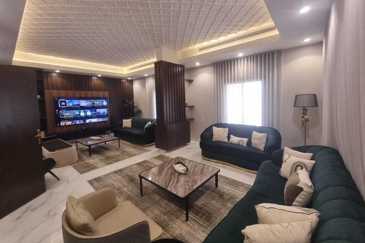 A modern living room with a flat-screen TV, dark sofas, a coffee table, and a large window with curtains.