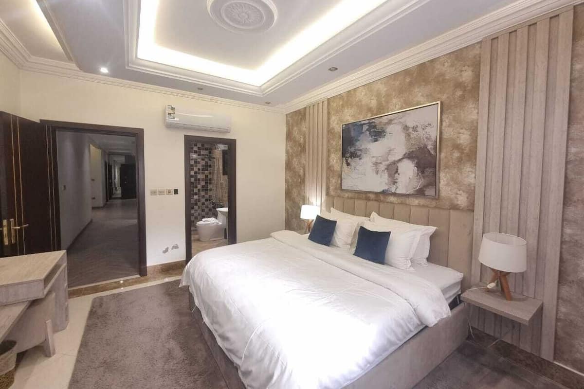 A spacious bedroom with a large bed, a mirror, a vanity, and a ceiling light fixture.