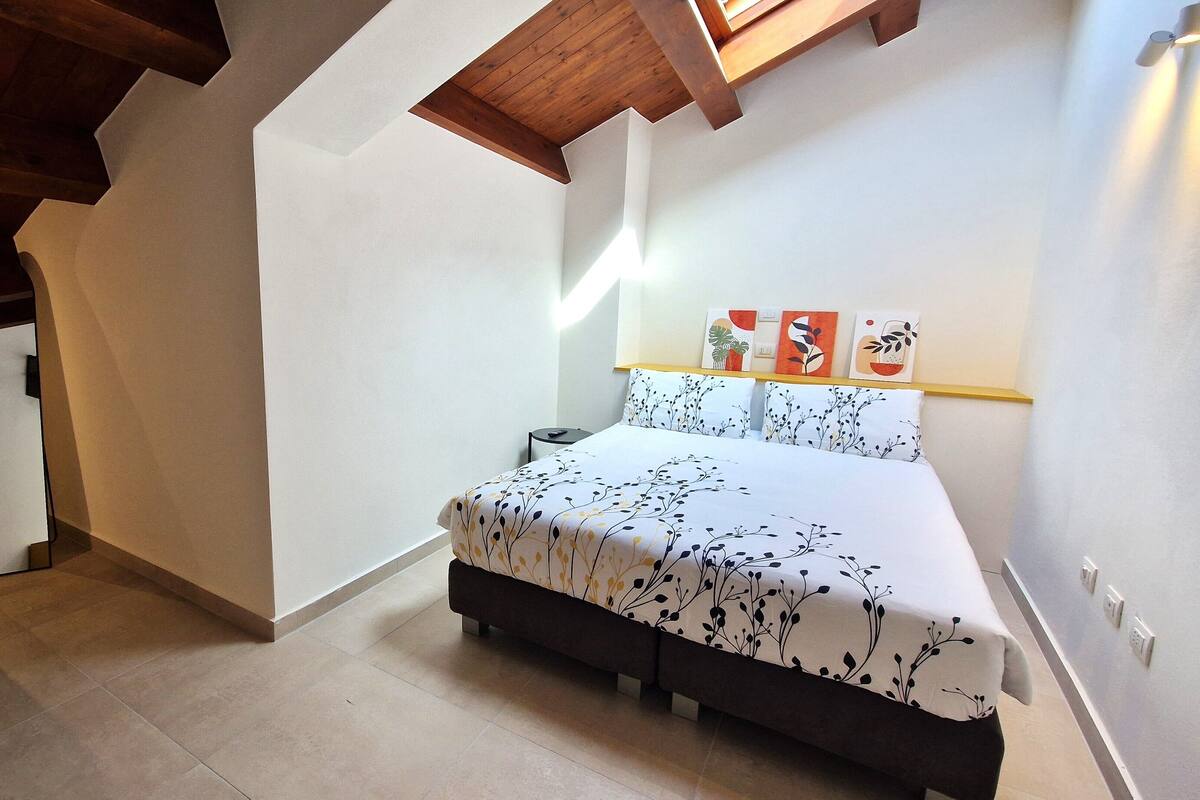 A bedroom with a bed, a nightstand, a wall-mounted shelf with decorative items, and a wooden ceiling beam.