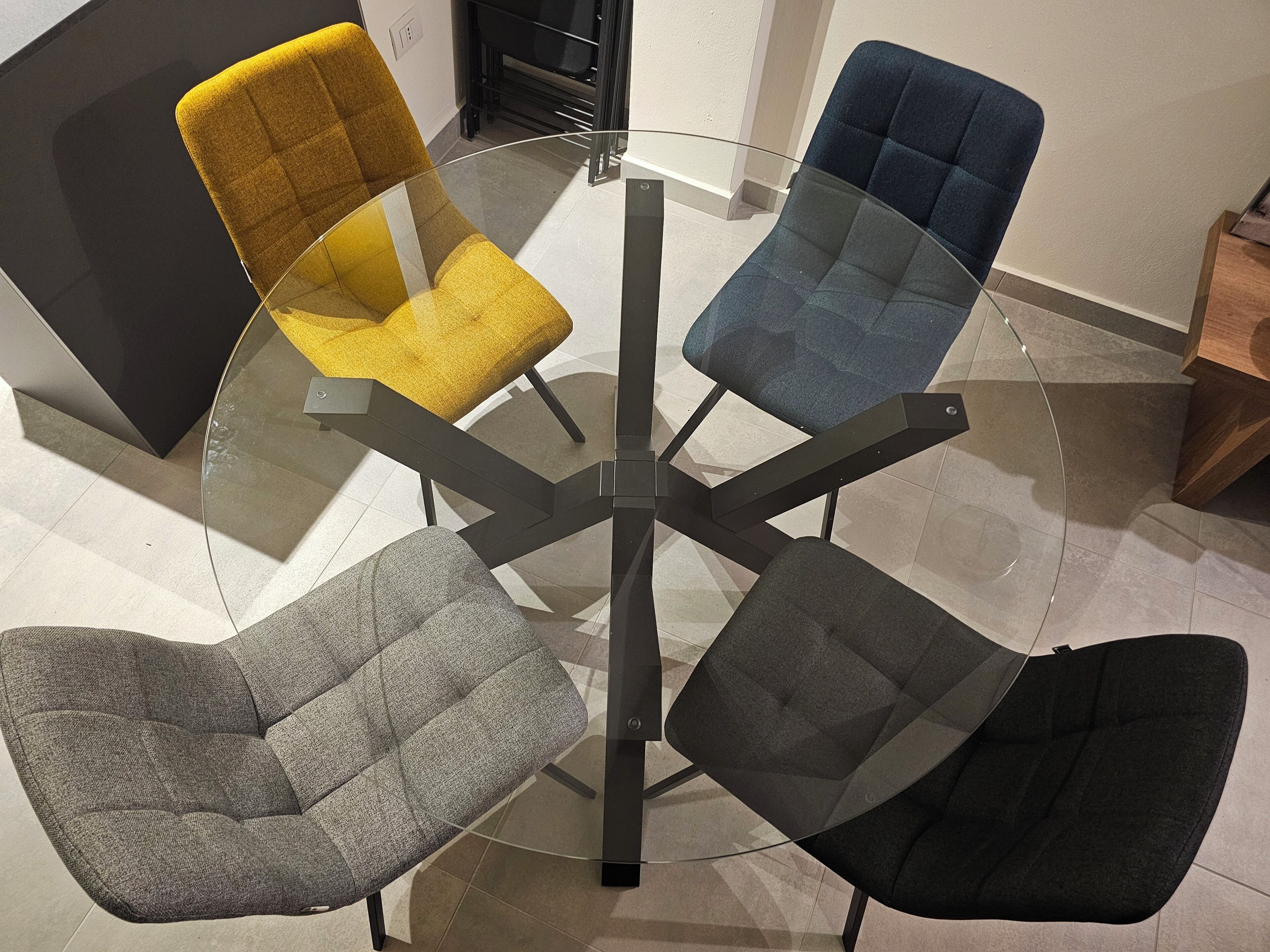 Una mesa redonda de vidrio rodeada por cuatro sillas de diferentes colores de tela.