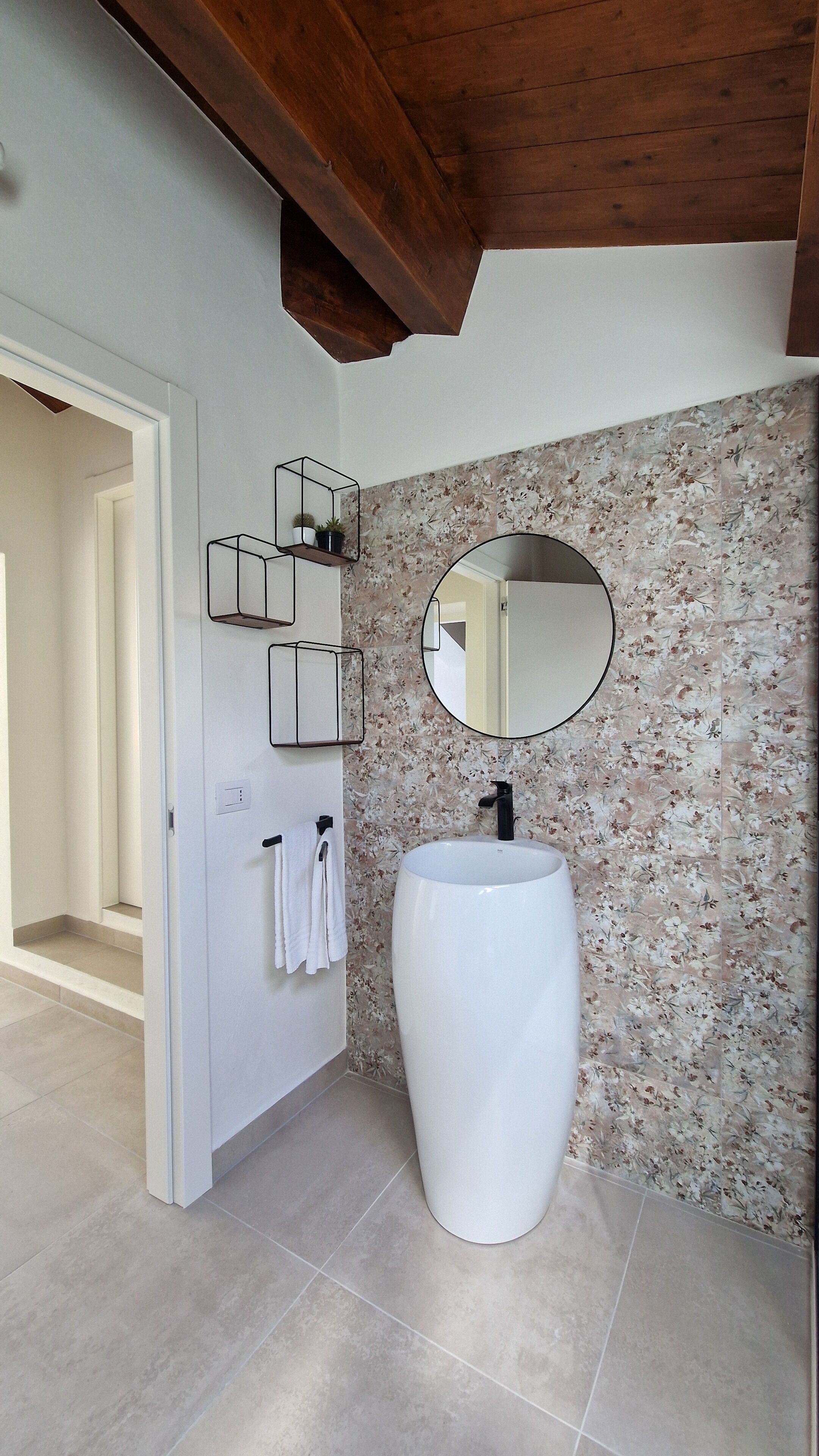 A bathroom with a white pedestal sink, a wooden ceiling, and a floral-patterned wall.