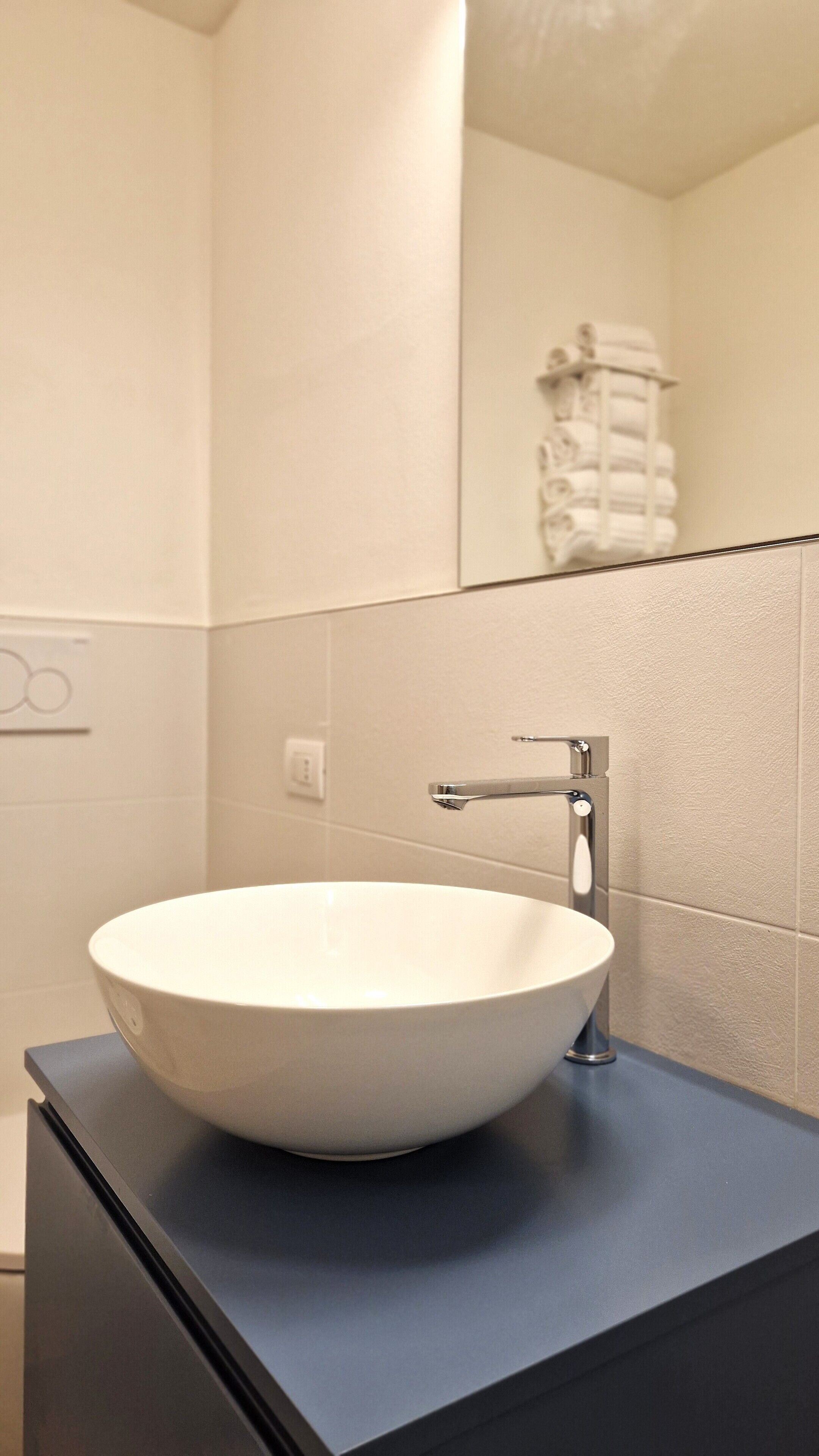 Un baño moderno con un lavamanos blanco tipo recipiente sobre una encimera oscura, un grifo cromado y un espejo que refleja un perchero.