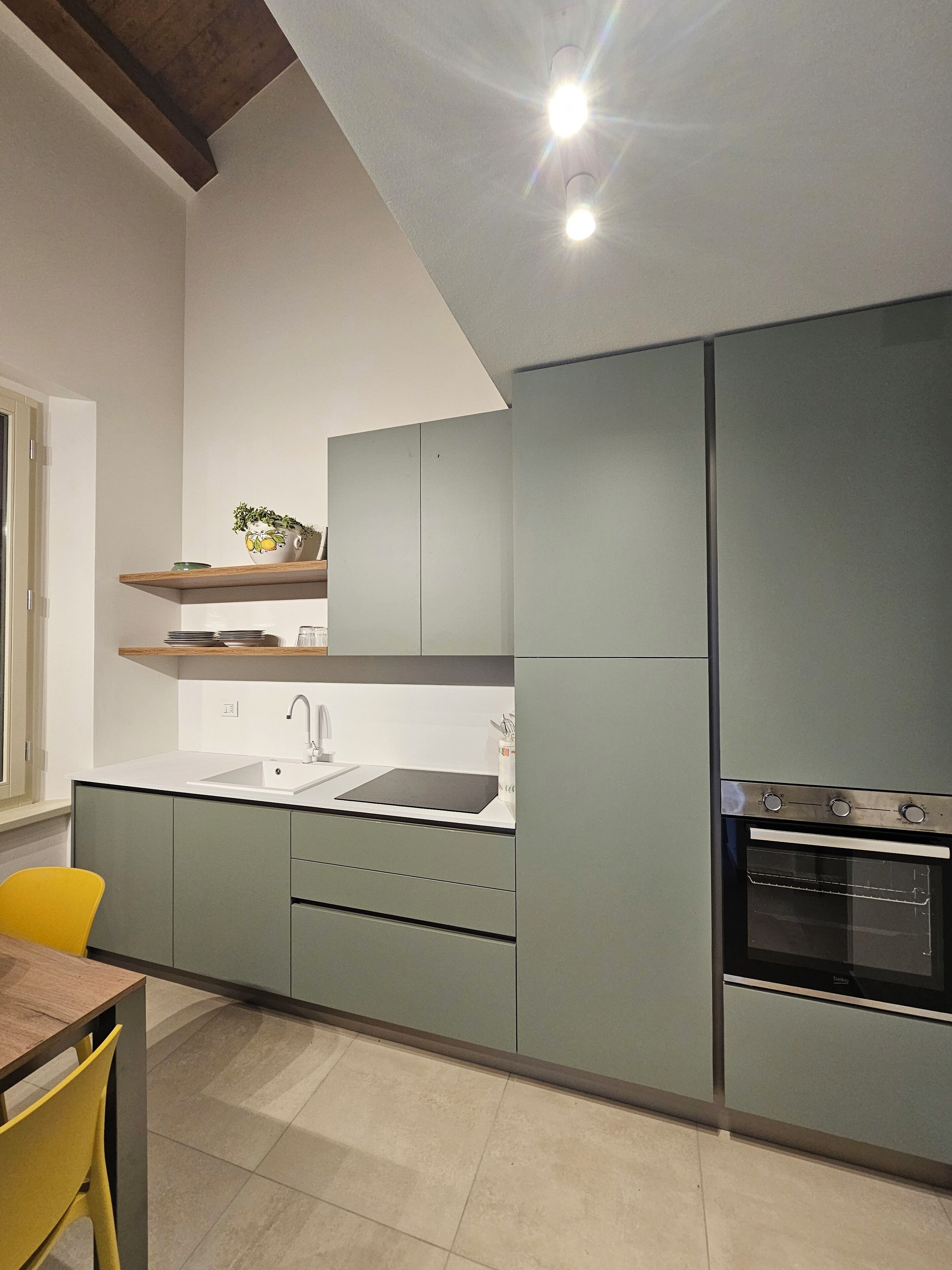Une cuisine moderne avec des meubles gris, un évier et une cuisinière. On y trouve une chaise jaune et une fenêtre sur le côté gauche.