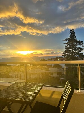 A balcony with a metal table and chair, overlooking a coastal landscape with a sunset and a prominent tree. - Modern 2BR Apartment in Caniço town w/ Pool, Sea Views & Parking – Near Funchal (Caniço)