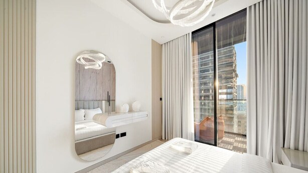 A modern bedroom with a large bed, a built-in vanity, a large mirror, and a balcony with a city view. - Timeless Luxury 1BR Apartment Near Dubai Marina & Iconic Palm Jumeirah (JVC)
