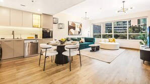 A modern apartment with a kitchen, dining area, and living room. It features a large window, a green sofa, and a white dining table.