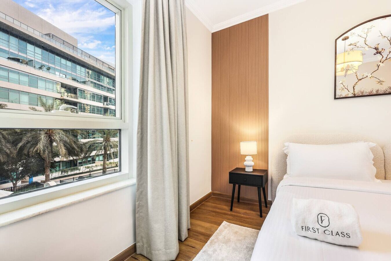 A hotel room with a large window, a bed with a 'First Class' pillow, a nightstand with a lamp, and a wooden panel wall.