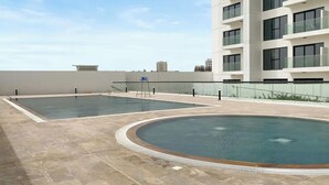 Ein Poolbereich auf dem Dach mit zwei rechteckigen Pools und einem kreisförmigen Pool, umgeben von einer niedrigen Mauer und einem modernen Gebäude mit mehreren Balkonen.