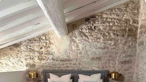A bedroom with a stone wall, a bed with grey bedding, two bedside tables with lamps, and a grey bench at the foot of the bed. - Duplex 2 minutes from the arena and maison carrée (Nîmes)