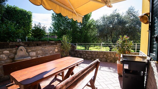 Een terras met een houten tafel en bank, een gele parasol en een stenen muur.