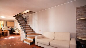 Een woonkamer met een beige bank, een houten trap en een keuken met houten kastjes en een betegelde achterwand.