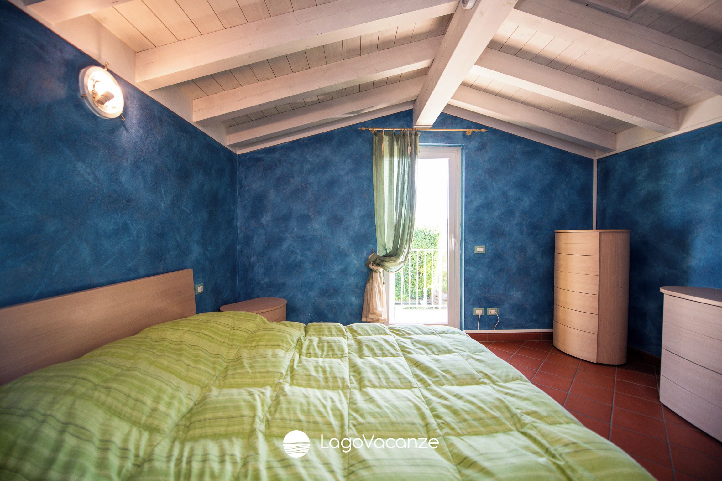 Una camera da letto con un letto, un comodino, una cassettiera, una finestra con tende e un soffitto con travi in legno.