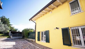 A yellow building with a stone-paved entrance, a gated driveway, and a window with shutters. - Casa Giardino (Polpenazze del Garda)