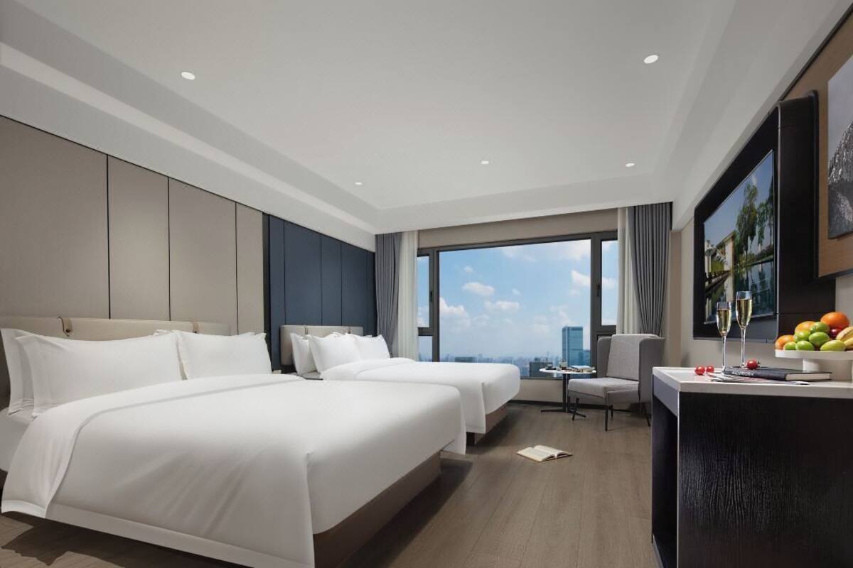 Une chambre d’hôtel avec deux lits, un bureau avec des fruits et des boissons, et une grande fenêtre donnant sur la ville.