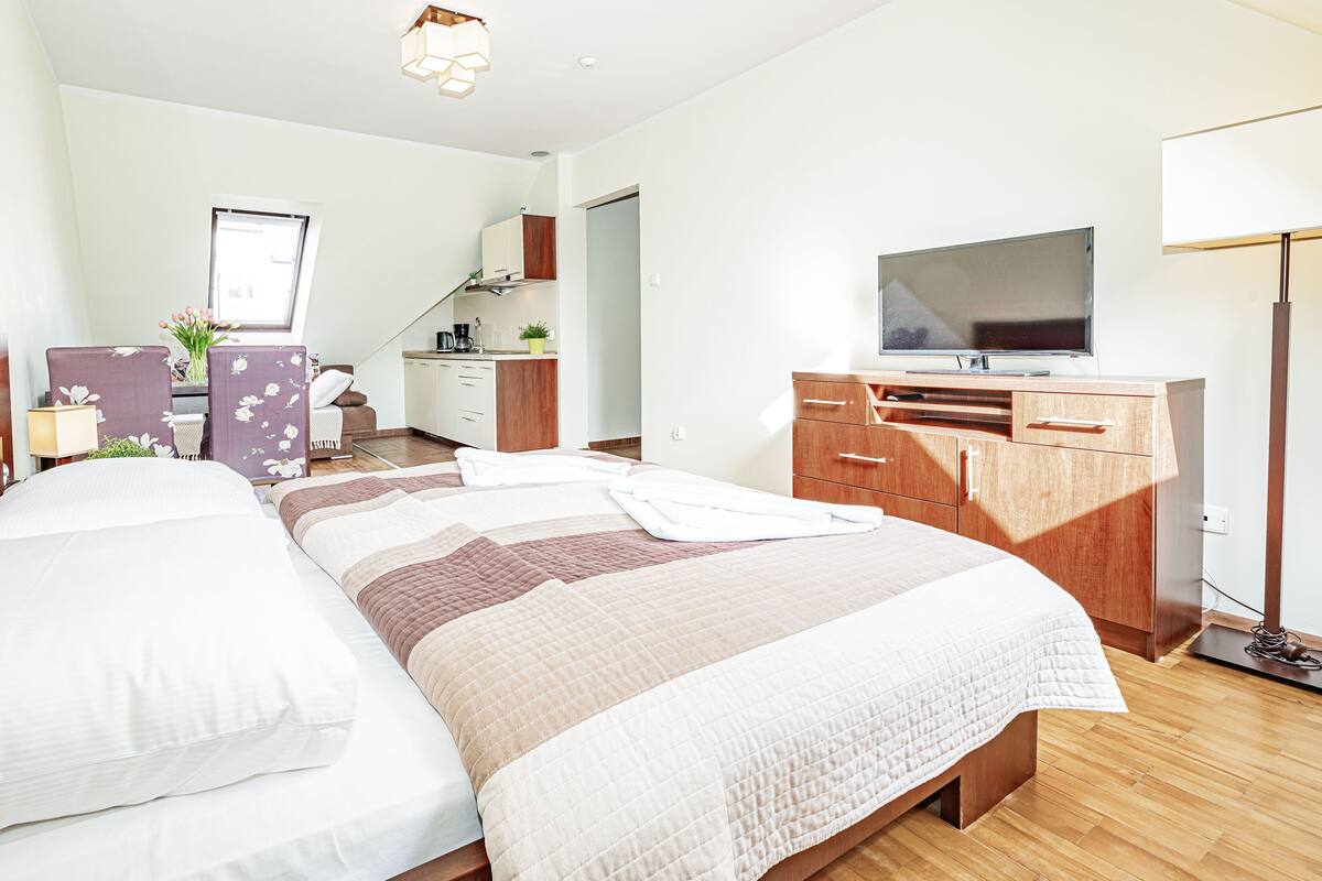 A hotel room with two beds, a TV, a desk with a chair, and a kitchenette in the background.