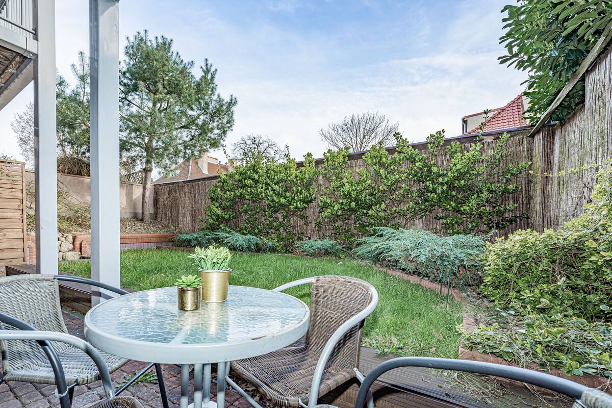 A patio with a round glass table and wicker chairs, surrounded by a wooden fence and lush greenery.