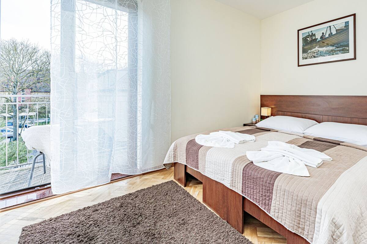 A hotel room with a large bed, a bedside table with a lamp, a window with sheer curtains, and a framed picture on the wall.