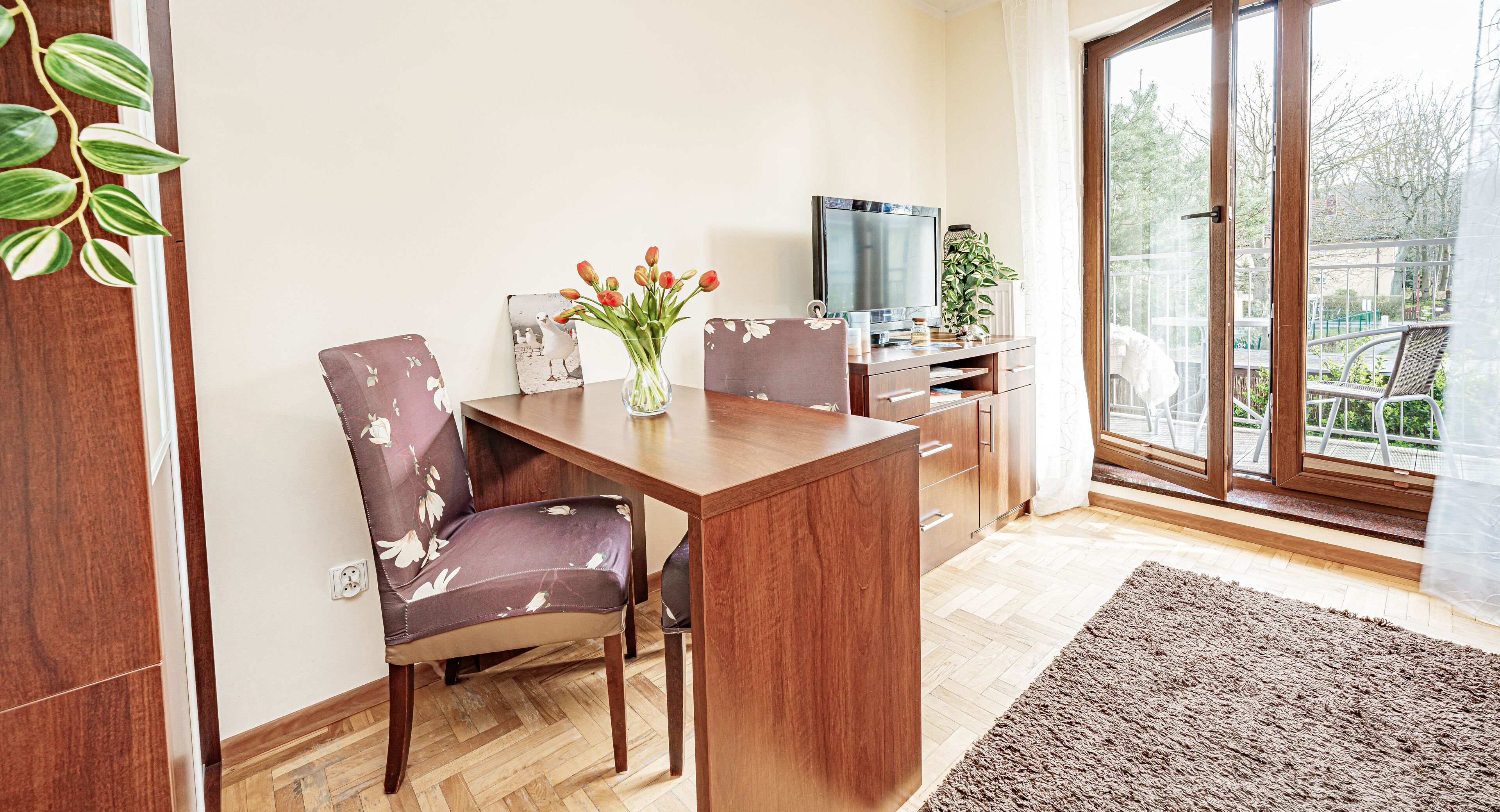 Ein moderner Essbereich mit einem Holztisch, zwei Stühlen, einer Vase mit Blumen, einem Fernseher und Blick auf einen Balkon mit Sitzmöbeln.