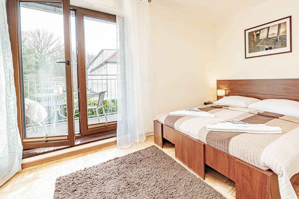 A hotel room with a large bed, a wooden headboard, a view through a sliding glass door, a framed picture on the wall, and a small table with a lamp.