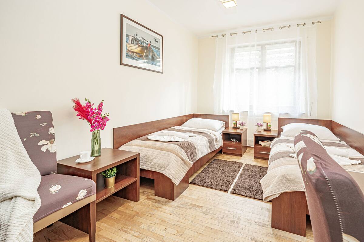 A hotel room with a bed, two bedside tables, a chair, a small table with a vase of flowers, and a window with sheer curtains.