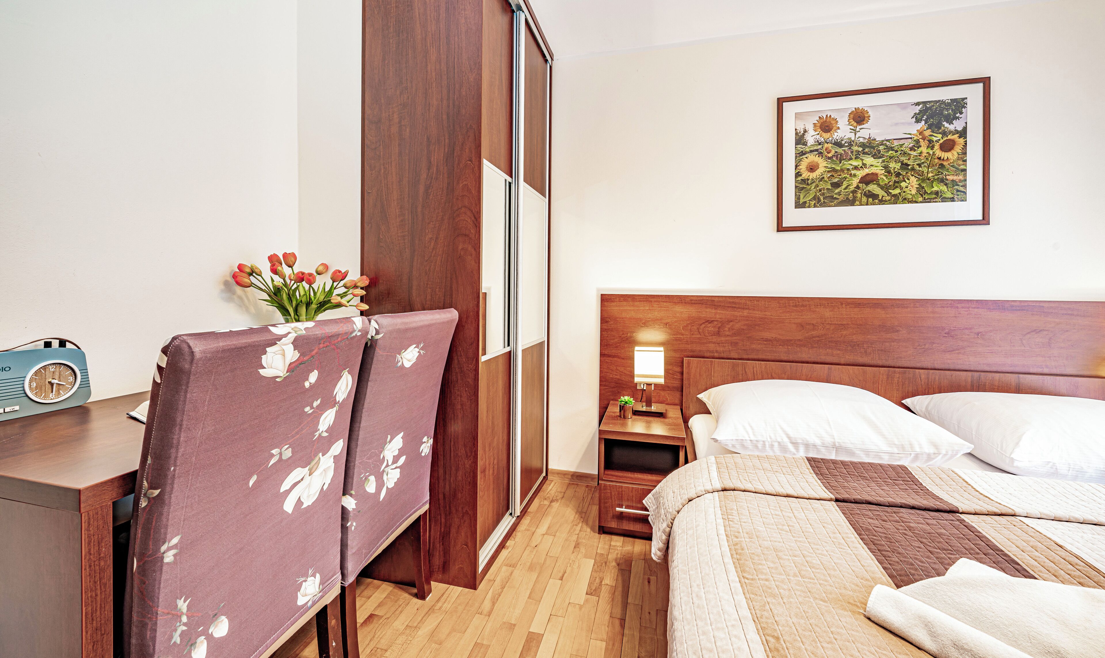 Habitación de hotel con cabecera de madera, cama con varias almohadas, mesita de noche con lámpara, planta pequeña y un cuadro en la pared.