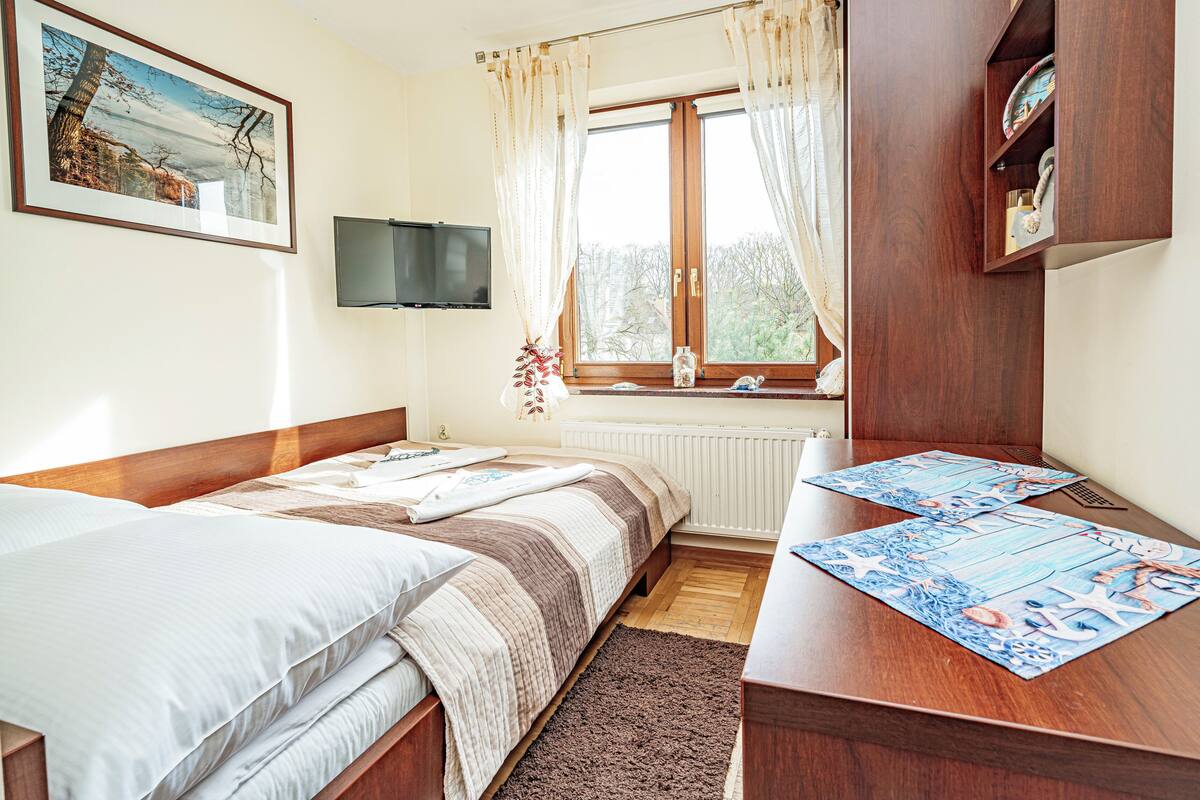A bedroom with two beds, a wooden desk, a window with curtains, a television on the wall, and a framed picture above the bed.