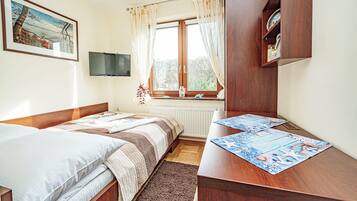 Une chambre avec deux lits, un bureau en bois, une fenêtre avec des rideaux, un téléviseur fixé au mur et un tableau encadré au-dessus du lit.