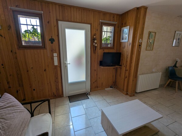 Quarto com painéis de madeira, porta branca com vidro jateado, televisão fixada na parede, uma mesa pequena e uma cadeira.