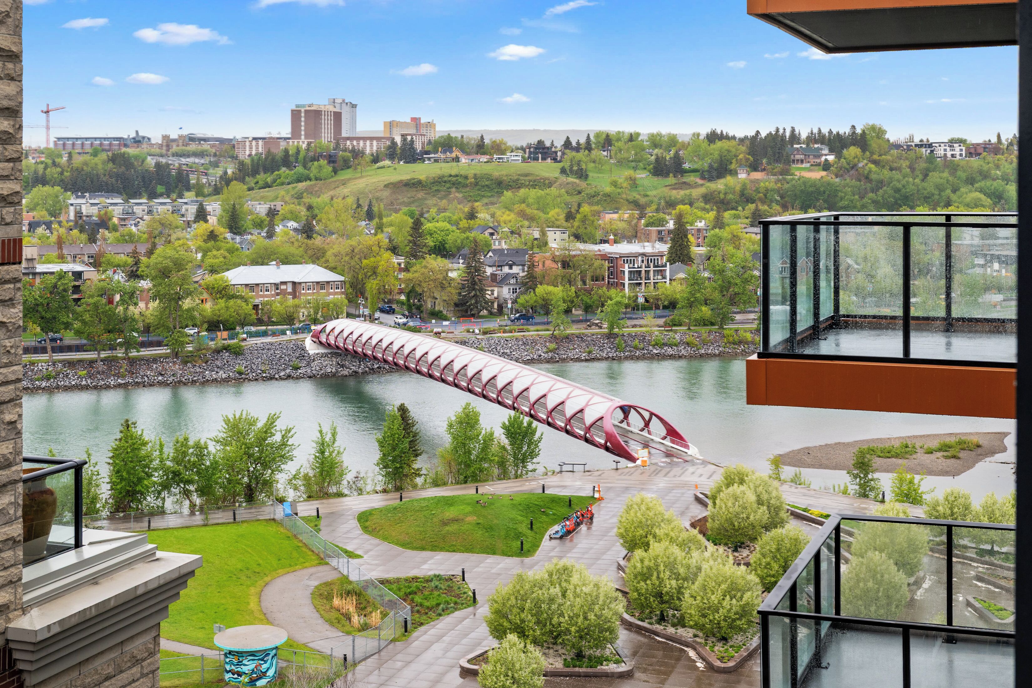 A view from a balcony overlooking a river with a red bridge, a park with a small lake, and buildings in the background.
