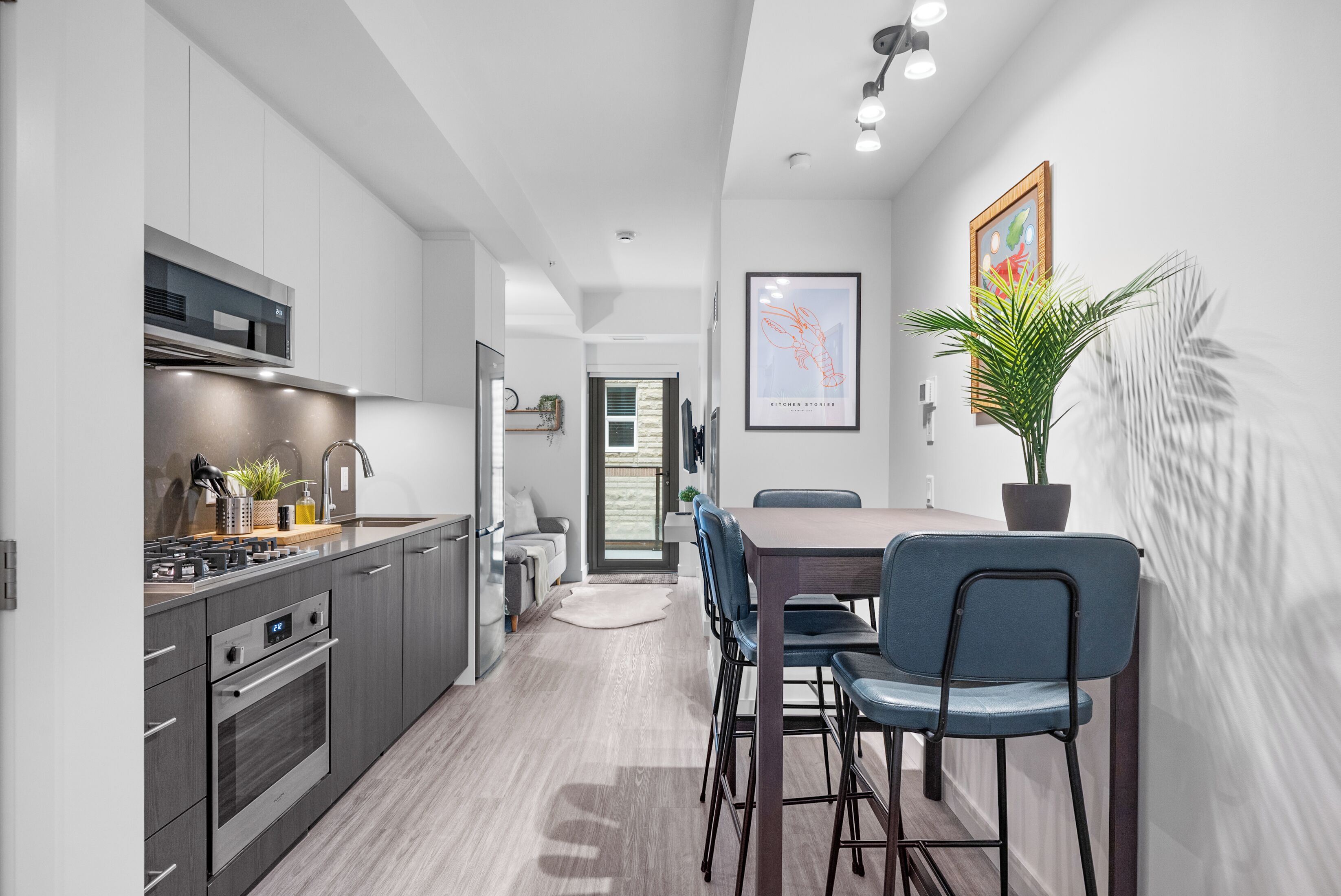 A modern kitchen with stainless steel appliances, a dining area with a wooden table and blue chairs, and a potted plant on the wall.
