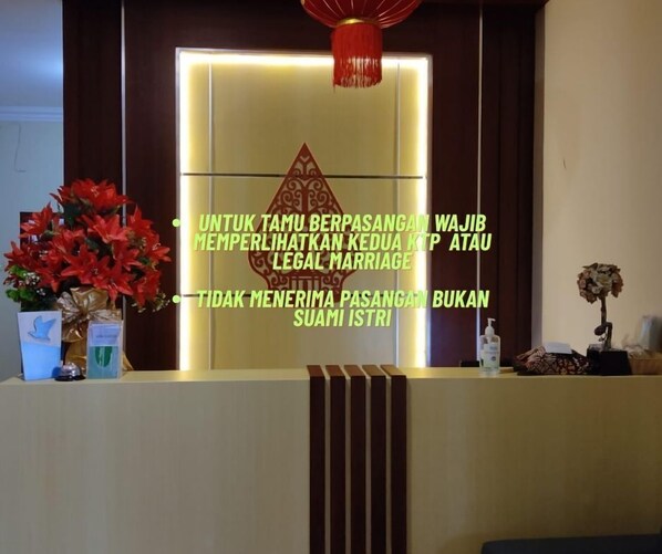 A reception desk with a sign in a foreign language, a red hanging ornament, and two flower arrangements. - Wisma Sejahtera Hotel (Magelang)