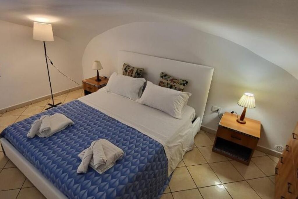 A neatly made bed with a blue quilt, two pillows, and two folded towels. There are two bedside tables, one with a lamp and the other with a small shelf. The room has a curved ceiling and tiled floor.