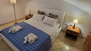 Una cama bien hecha con colcha azul, dos almohadas y dos toallas dobladas. Hay dos mesitas de noche, una con una lámpara y la otra con una pequeña estantería. La habitación tiene techo curvo y suelo de baldosas.