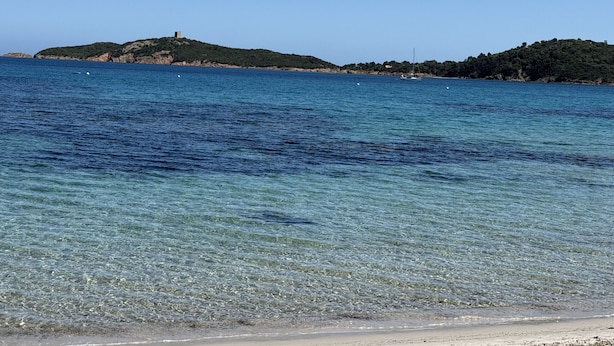 Eine Küstenlandschaft mit klarem, blauem Wasser, einem Sandstrand und einem Hügel mit einer Bebauung.