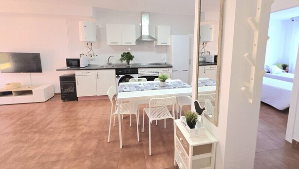 Een moderne keuken met witte kasten, een eettafel met stoelen, een televisie en een grote spiegel.