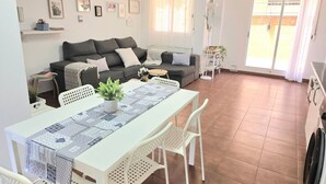 Een ruime woonkamer met een eettafel, witte stoelen, een grijze bank en een keukenhoek.
