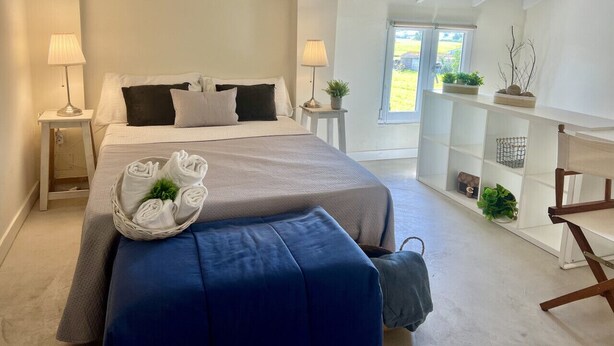 Een slaapkamer met een groot bed, bedlampen, een klein tafeltje, een bankje en een raam met uitzicht op de omgeving.