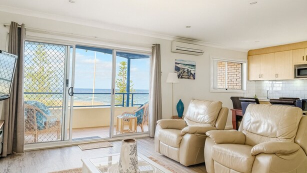 A living room with a view of the ocean, a balcony with a chair, and a kitchen area. - Amazing Ocean Views, Spacious Waterfront Holiday Apartment (The Entrance)
