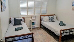 A bedroom with two single beds, a ceiling fan, a window with blinds, and a small bedside table with a vase.