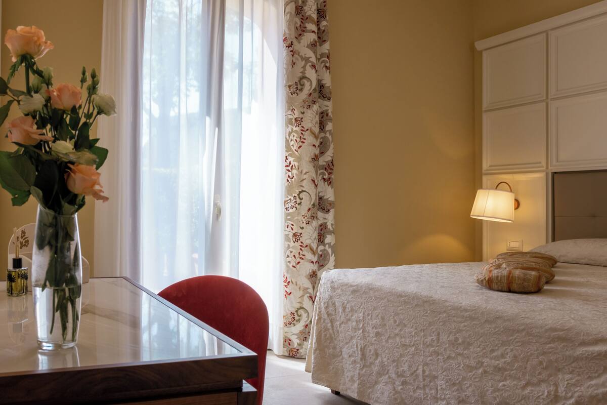A hotel room with a bed, a bedside table with a lamp, a vase with flowers, and a chair.