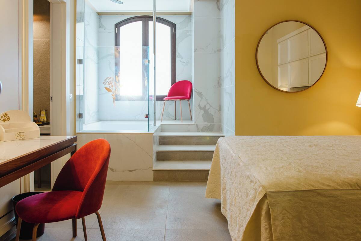 A hotel room with a bed, a desk, a chair, a mirror, and a view of a staircase and a door.