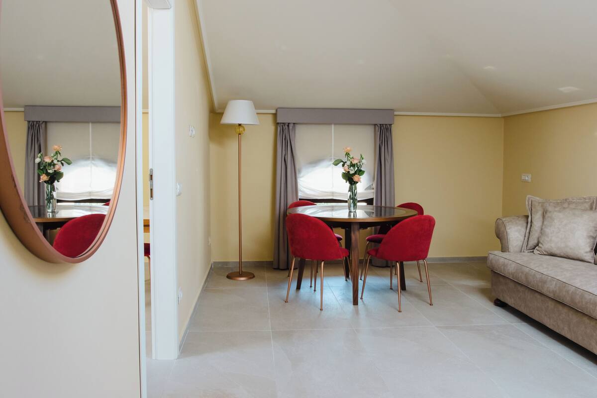 A modern living room with a beige sofa, a dining table with red chairs, and a large mirror on the wall.